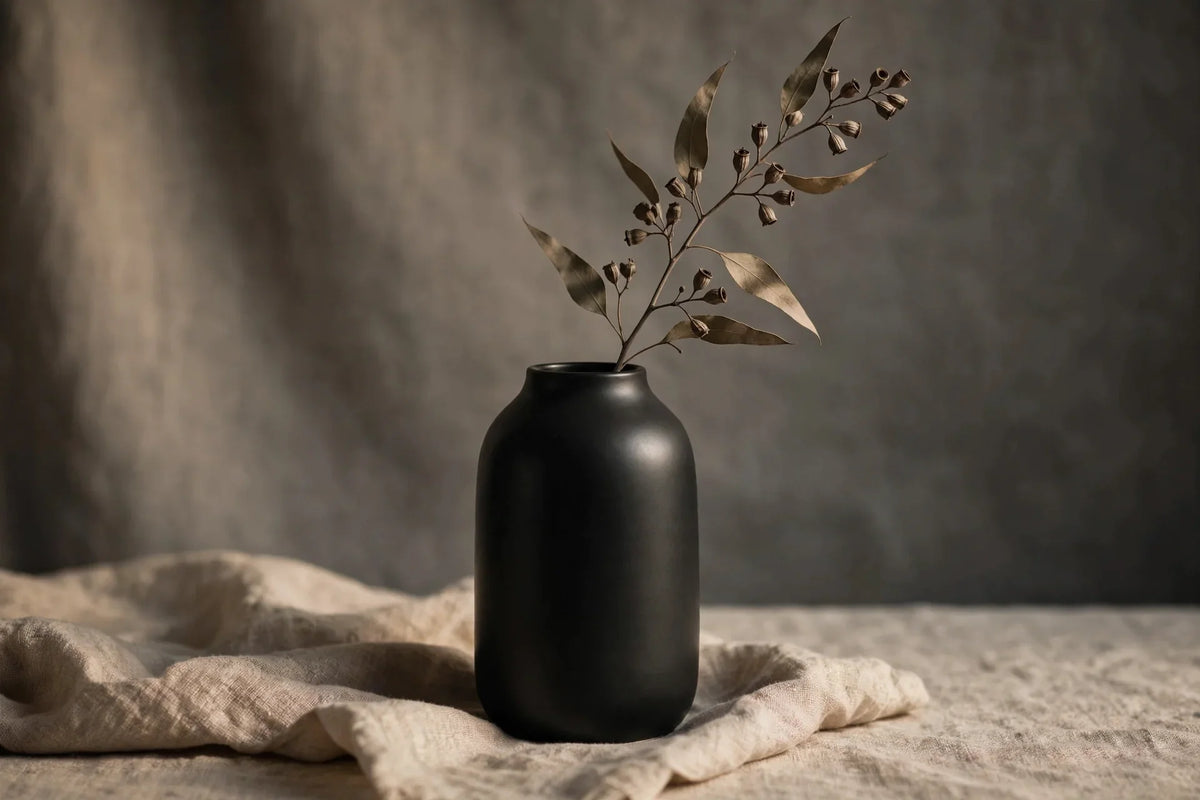 Matte black ceramic vase with dried eucalyptus on a textured linen surface, warm studio lighting